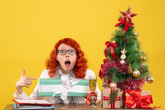 Vista frontal doctora sentada con regalos de Navidad y árbol sobre fondo amarillo