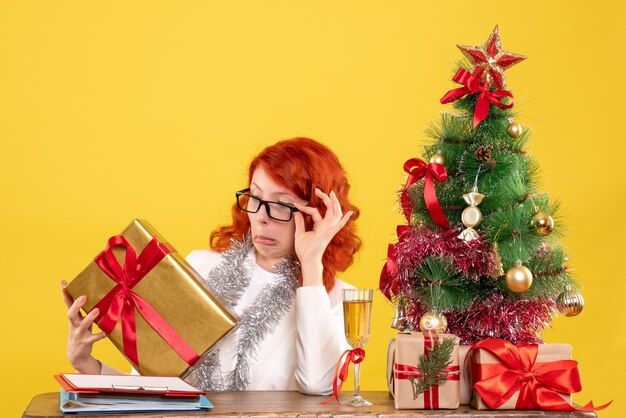 Vista frontal doctora sentada con regalos de Navidad y árbol sobre fondo amarillo