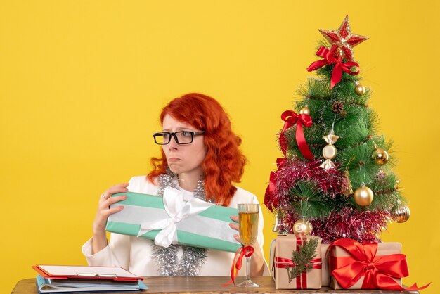 Vista frontal doctora sentada con regalos de Navidad y árbol sobre fondo amarillo