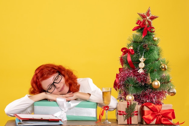 Vista frontal doctora sentada con regalos de Navidad y árbol sobre fondo amarillo