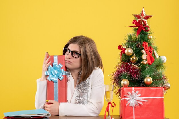 Vista frontal doctora sentada con regalos de Navidad y árbol sobre fondo amarillo