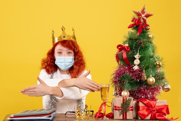 Vista frontal de la doctora sentada con regalos de Navidad y con corona sobre fondo amarillo