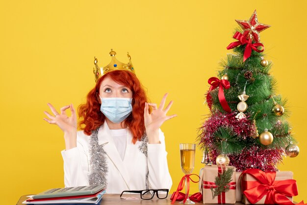 Vista frontal de la doctora sentada con regalos de Navidad y con corona sobre fondo amarillo