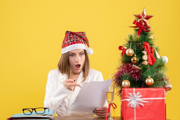 Vista frontal doctora sentada con regalos de Navidad leyendo documentos sobre fondo amarillo