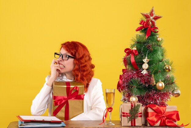 Vista frontal doctora sentada con regalos de Navidad sobre fondo amarillo