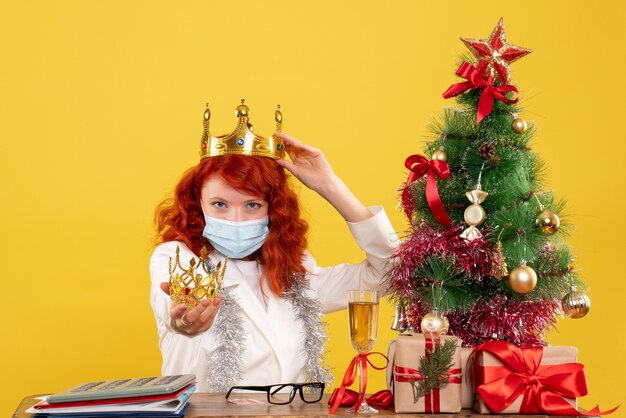 Vista frontal doctora sentada con regalos de Navidad y sosteniendo la corona en el escritorio amarillo