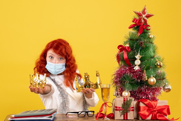 Vista frontal doctora sentada con regalos de Navidad y sosteniendo coronas sobre fondo amarillo