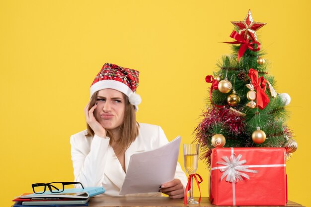 Vista frontal doctora sentada con regalos de Navidad sosteniendo documentos aburridos sobre fondo amarillo