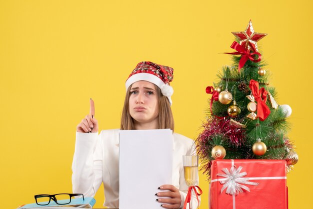 Vista frontal doctora sentada con regalos de Navidad sosteniendo documentos en el escritorio amarillo