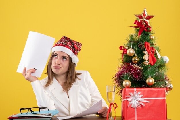 Vista frontal doctora sentada con regalos de Navidad sosteniendo documentos sobre el fondo amarillo