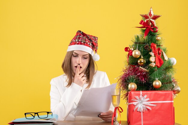 Vista frontal doctora sentada con regalos de Navidad sosteniendo documentos sobre el fondo amarillo