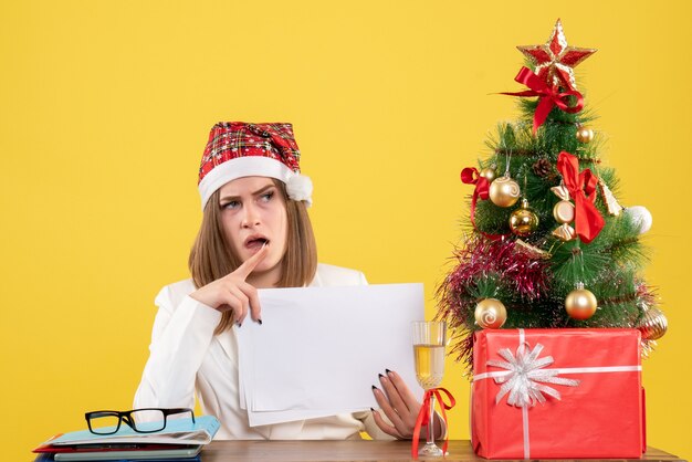 Vista frontal doctora sentada con regalos de Navidad sosteniendo documentos sobre el fondo amarillo