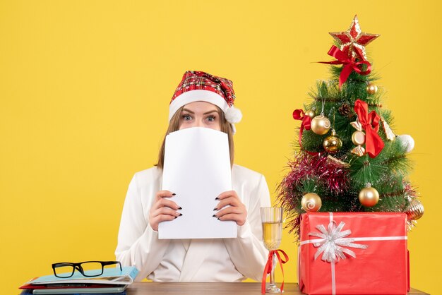 Vista frontal doctora sentada con regalos de Navidad sosteniendo documentos sobre el fondo amarillo