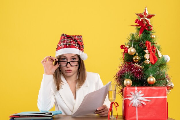 Vista frontal doctora sentada con regalos de Navidad sosteniendo documentos sobre el fondo amarillo