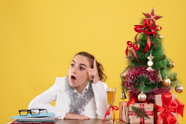 Vista frontal doctora sentados alrededor de regalos de Navidad y árbol sobre fondo amarillo