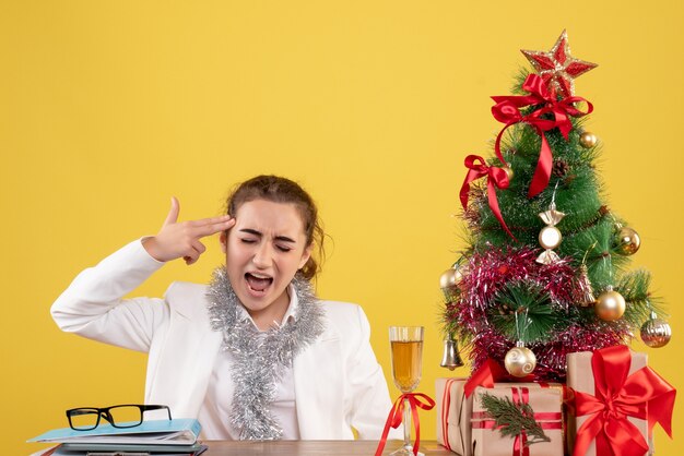 Vista frontal doctora sentados alrededor de regalos de Navidad y árbol sobre fondo amarillo