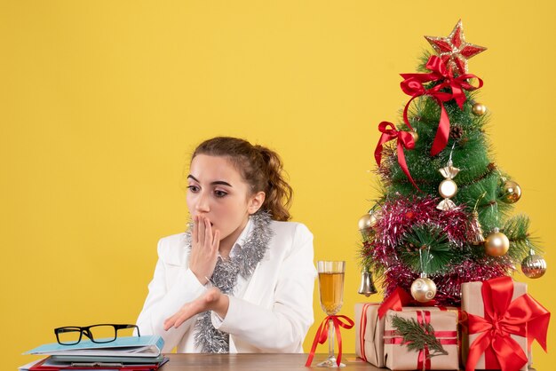 Vista frontal doctora sentados alrededor de regalos de Navidad y árbol sobre fondo amarillo