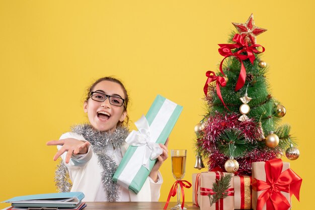 Vista frontal doctora sentados alrededor de regalos de Navidad y árbol sobre fondo amarillo