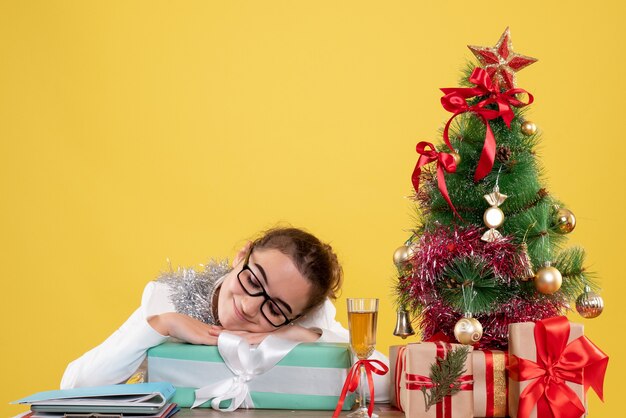 Vista frontal doctora sentados alrededor de regalos de Navidad y árbol sobre fondo amarillo