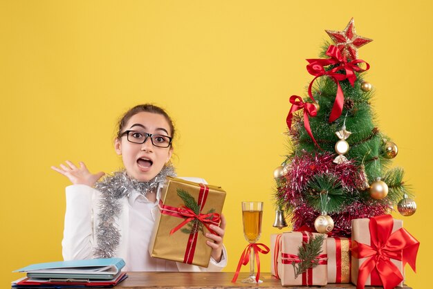 Vista frontal doctora sentados alrededor de regalos de Navidad y árbol sobre fondo amarillo