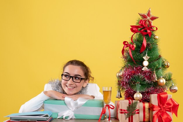 Foto gratuita vista frontal doctora sentados alrededor de regalos de navidad y árbol sonriendo sobre fondo amarillo