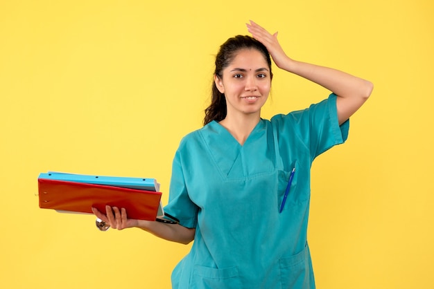 Vista frontal de la doctora sonrió en uniforme sosteniendo carpetas de pie en la pared amarilla