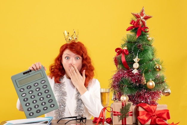 Vista frontal doctora sosteniendo la calculadora alrededor de regalos de Navidad y árbol