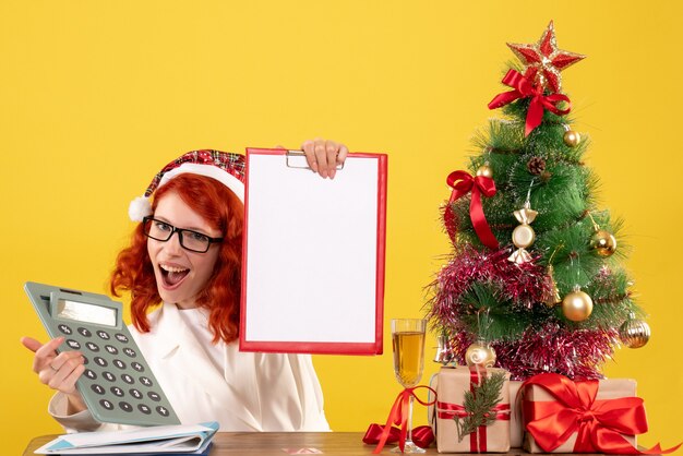 Vista frontal doctora sosteniendo la calculadora alrededor de regalos de Navidad y árbol