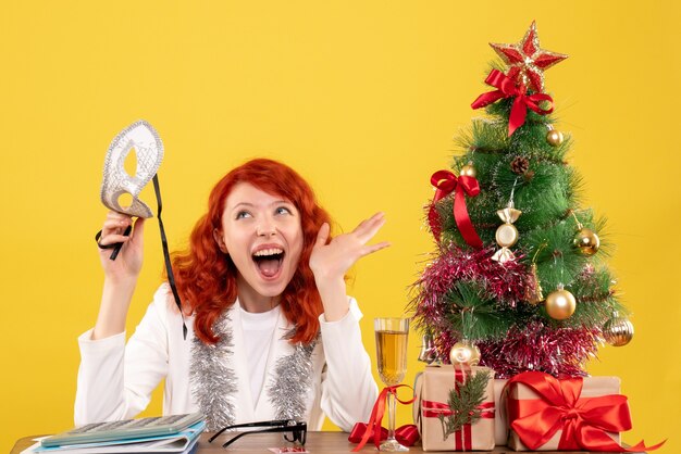 Vista frontal doctora sosteniendo la máscara alrededor del árbol de Navidad y regalos