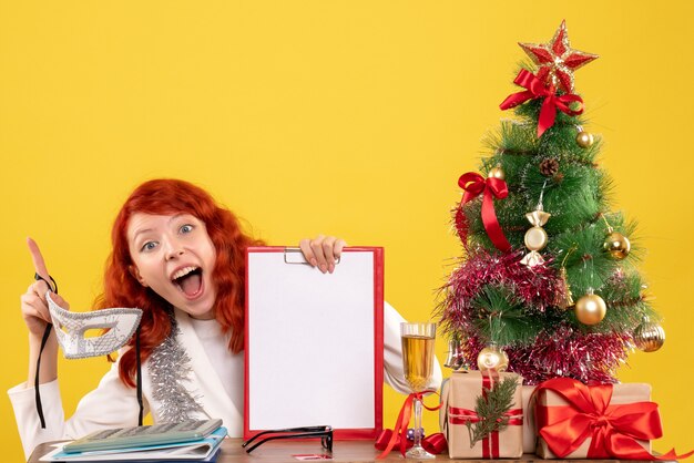 Vista frontal doctora sosteniendo máscara y nota de archivo alrededor del árbol de Navidad