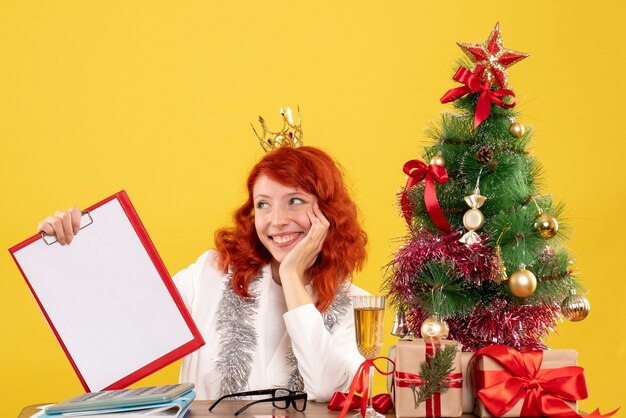 Vista frontal doctora sosteniendo la nota de archivo alrededor del árbol de Navidad y regalos