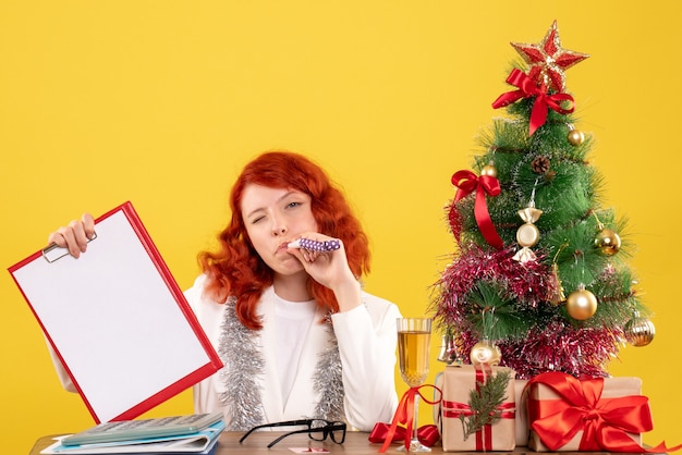 Vista frontal doctora sosteniendo la nota de archivo alrededor del árbol de Navidad y regalos