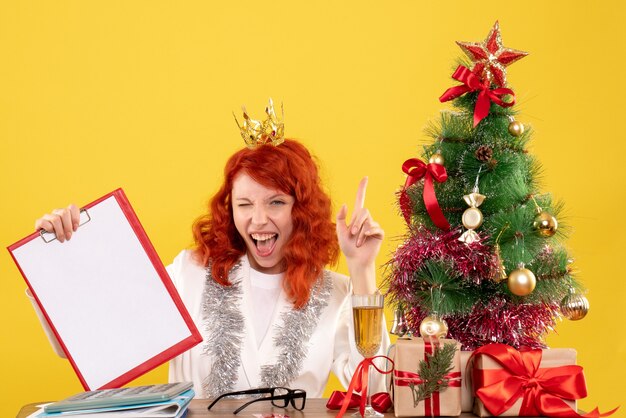 Vista frontal doctora sosteniendo la nota de archivo alrededor del árbol de Navidad y regalos