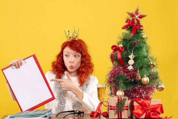 Vista frontal doctora sosteniendo la nota de archivo alrededor del árbol de Navidad y regalos