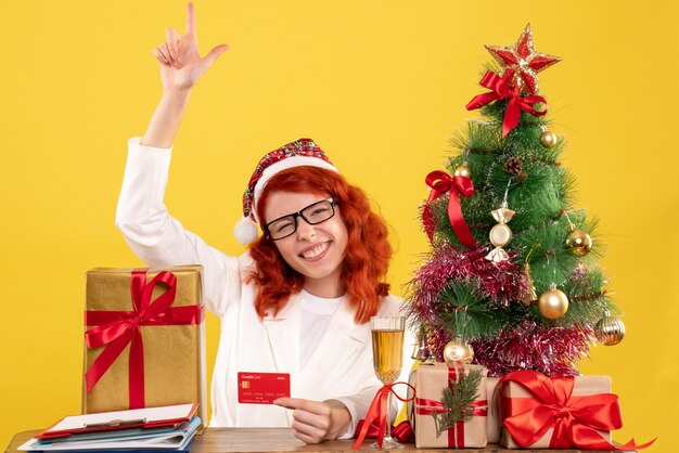 Vista frontal doctora sosteniendo tarjeta bancaria alrededor de árboles y regalos de Navidad