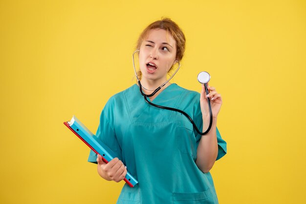 Vista frontal de la doctora en traje médico con análisis y estetoscopio en pared amarilla