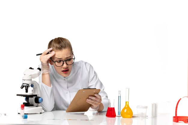 Vista frontal doctora en traje médico blanco escribiendo notas sobre un espacio en blanco claro