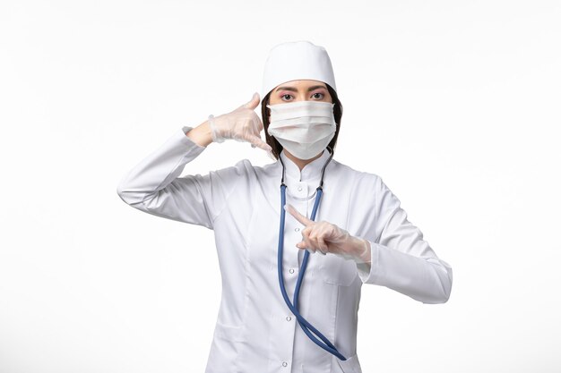 Vista frontal doctora en traje médico blanco estéril con máscara debido a coronavirus en pared blanca clara enfermedad enfermedad pandémica covid-virus