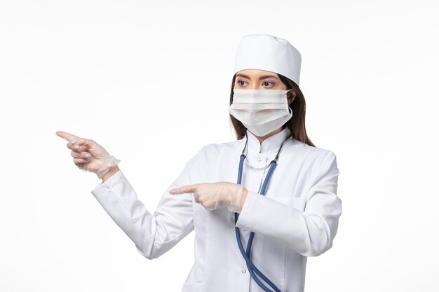 Vista frontal doctora con traje médico blanco estéril con máscara debido a coronavirus en la pared blanca enfermedad covid-enfermedad del virus pandémico