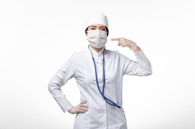 Vista frontal doctora en traje médico blanco estéril con máscara debido a covid- en la pared blanca enfermedad covid- enfermedad pandémica