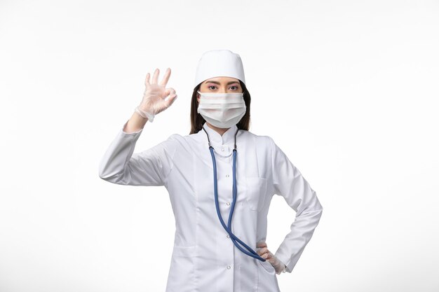 Vista frontal doctora en traje médico blanco estéril con máscara debido a covid- en la pared blanca enfermedad enfermedad por virus pandémico