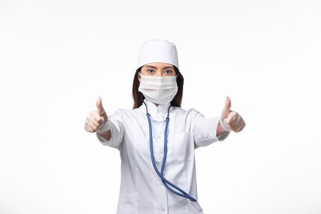 Vista frontal doctora en traje médico blanco estéril con máscara debido a covid- posando en la pared blanca enfermedad covid- enfermedad por virus pandémico
