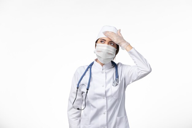Vista frontal de la doctora en traje médico blanco estéril y con máscara debido a covid- que tiene dolor de cabeza en la pared blanca enfermedad virus covid- enfermedad pandémica