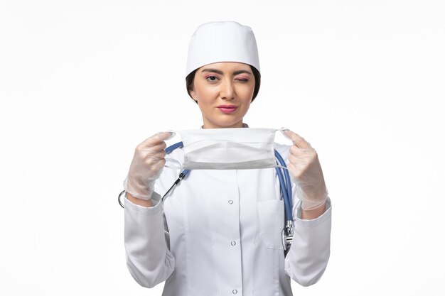 Vista frontal doctora en traje médico blanco estéril y sosteniendo una máscara debido a covid- en la pared blanca enfermedad covid- enfermedad por virus pandémico