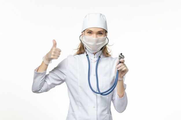 Vista frontal doctora en traje médico blanco con estetoscopio y sonriendo en el escritorio blanco enfermedad covid-enfermedad pandémica