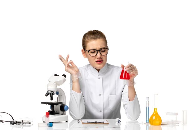 Vista frontal doctora en traje médico blanco con guantes sosteniendo matraz con solución en un escritorio blanco