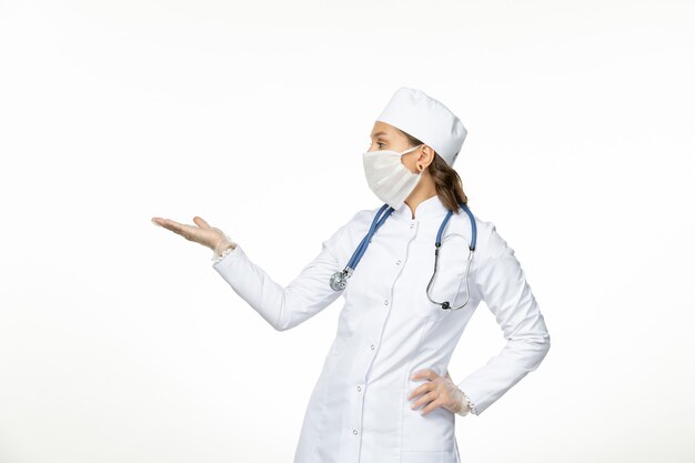 Vista frontal doctora en traje médico blanco con máscara debido al coronavirus en el aislamiento de la enfermedad pandémica de pared blanca clara