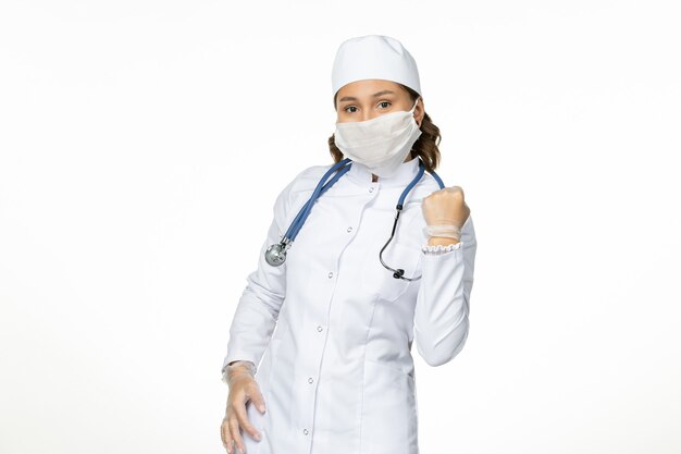 Vista frontal doctora en traje médico blanco y con máscara debido al regocijo del coronavirus en la pared blanca aislamiento de la enfermedad pandémica covid