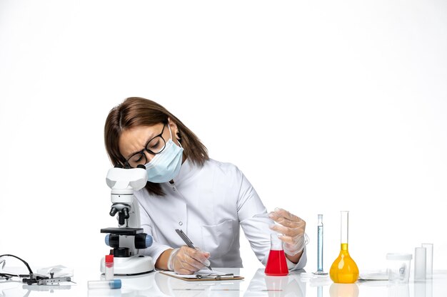 Vista frontal doctora en traje médico blanco con máscara debido a covid escribiendo notas en el espacio en blanco