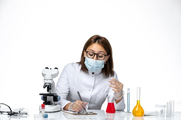 Vista frontal doctora en traje médico blanco con máscara debido a covid escribiendo notas en el espacio en blanco